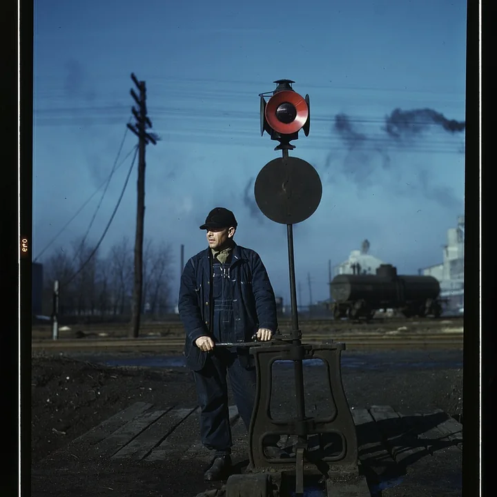 A railroad worker operates a track switch with a signal and rail yard behind him.