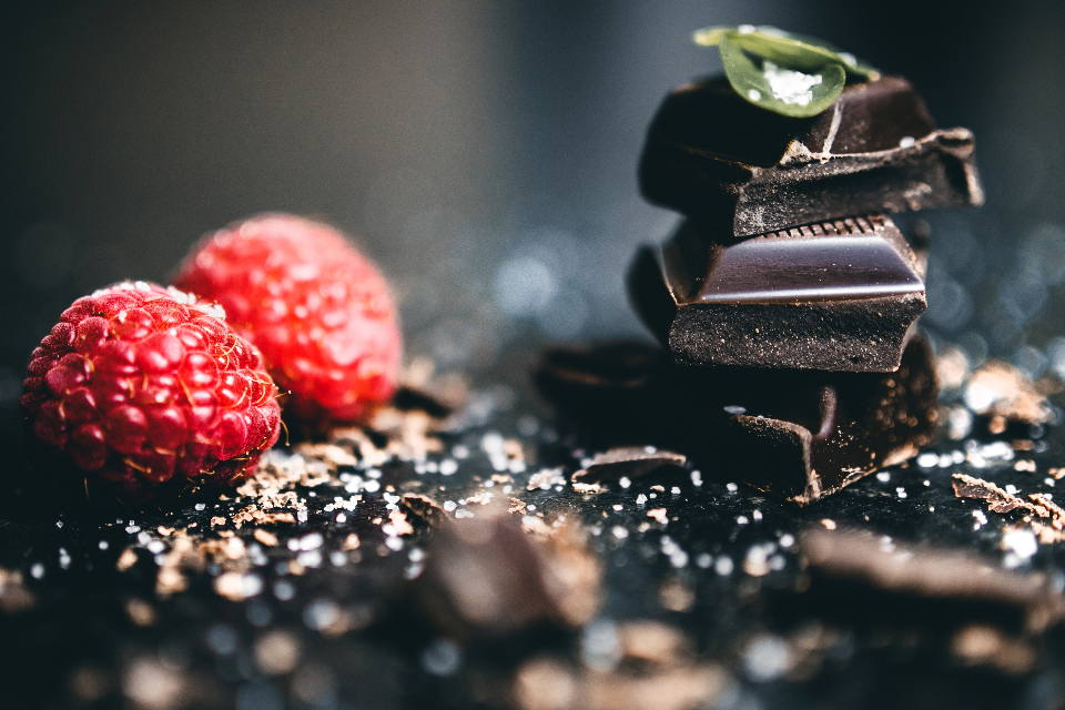 Pieces of dark chocolate stacked beside raspberries on a dark surface.