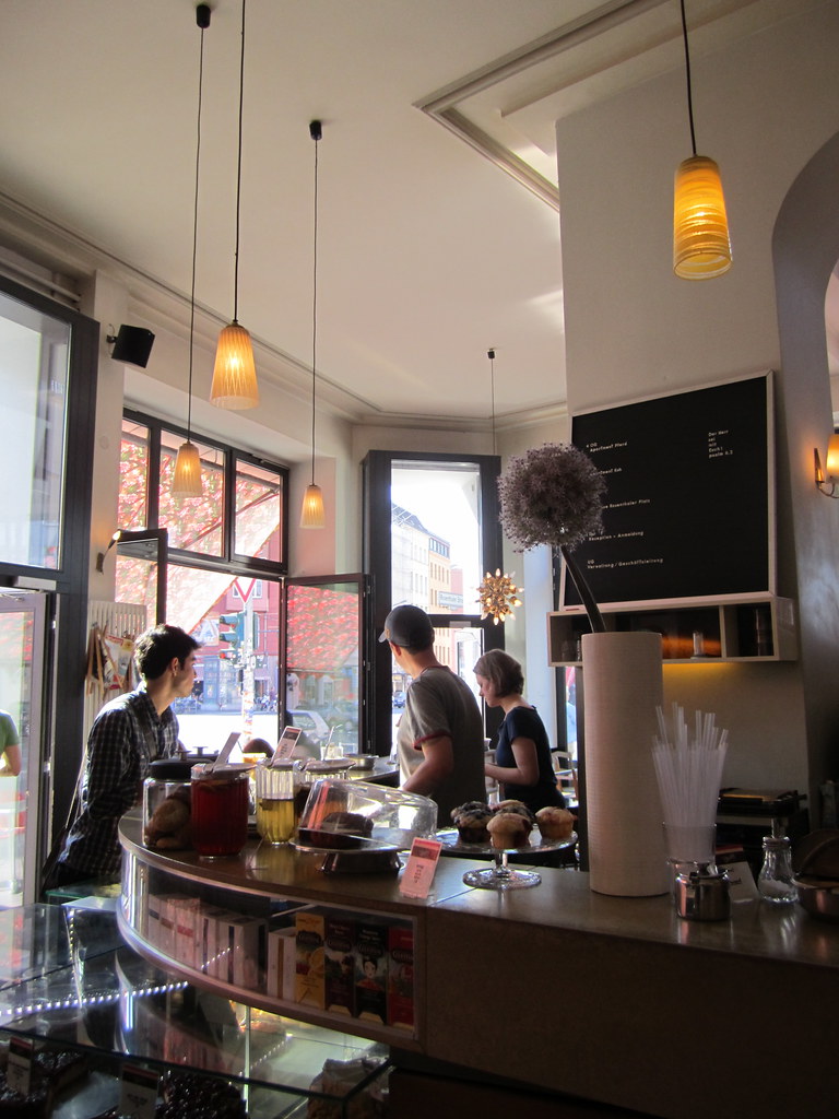 Light falls across a coffeehouse counter, cups, and people gathered around it.